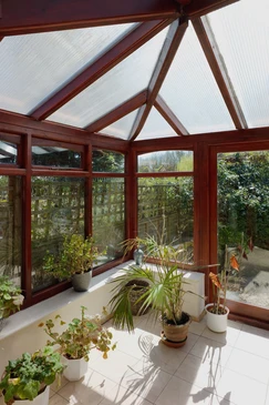 Véranda moderne avec structure en bois et toiture vitrée, aménagée en jardin d'hiver avec nombreuses plantes en pots disposées au sol et sur rebords